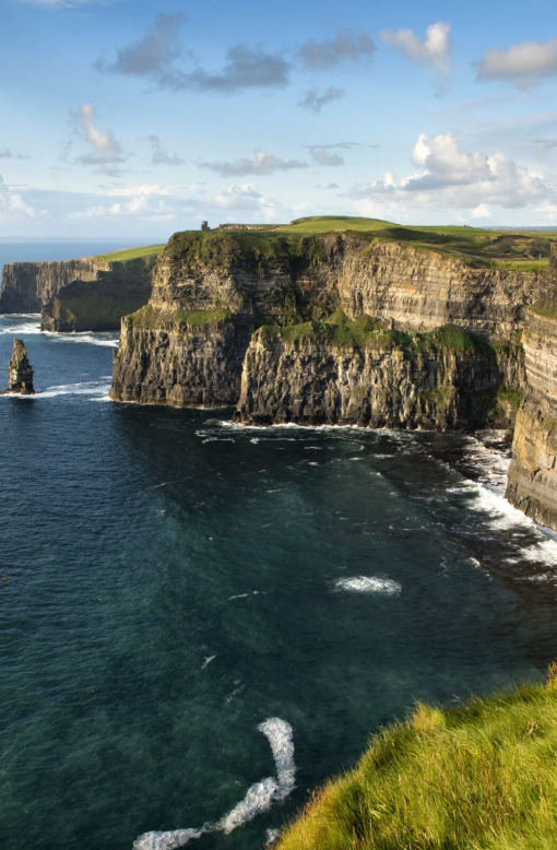 Magical view of the Cliffs of Moher towering over the Atlantic Ocean with waves crashing below on a summer day showing blue skies with minimal clouds.