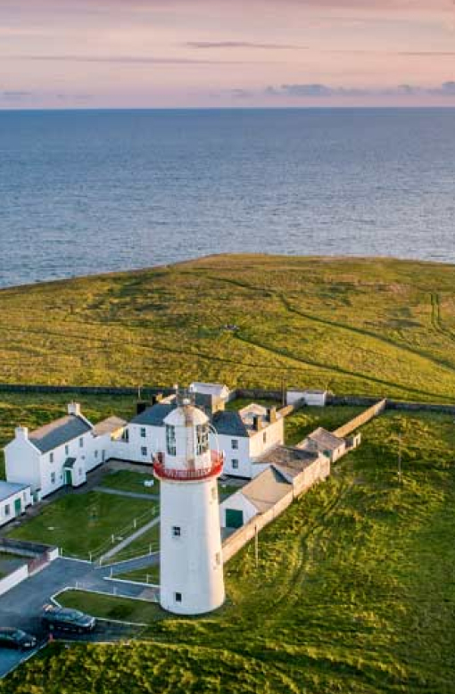 A stunning aerial view of Loop Head Lighthouse perched on a dramatic cliff edge, surrounded by the deep blue waters of the Atlantic Ocean with the sky painted pink by the evening sunset.