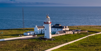 A stunning aerial view of Loop Head Lighthouse perched on a dramatic cliff edge, surrounded by the deep blue waters of the Atlantic Ocean.